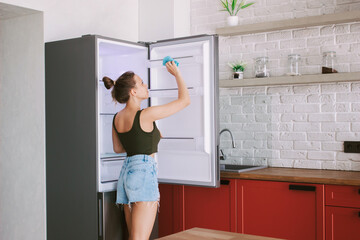 A young сaucasian woman cleans the refrigerator with a rag in the kitchen. Household chores,...