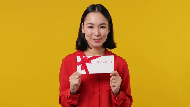 Surprised shocked happy young woman of Asian ethnicity 20s wear red shirt pointing finger on gift certificate voucher for store doing winner gesture isolated on plain yellow background studio portrait