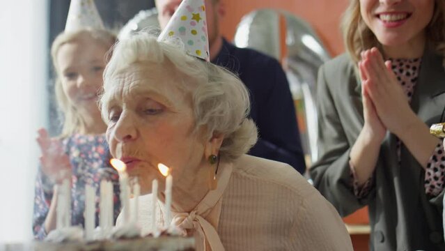 Joyous family in party hats hugging elderly grandmother after she blowing candles on birthday cake during home celebration