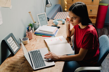 Young hispanic woman writing down notes while working with laptop