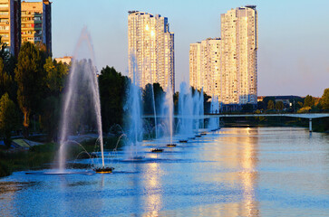Obraz premium Picturesque sunset landscape of Rusanivka neighborhood at sunny day. Fountains in the city channel. Modern skyscrapers in the background. Landscape during the autumn season. Kyiv, Ukraine