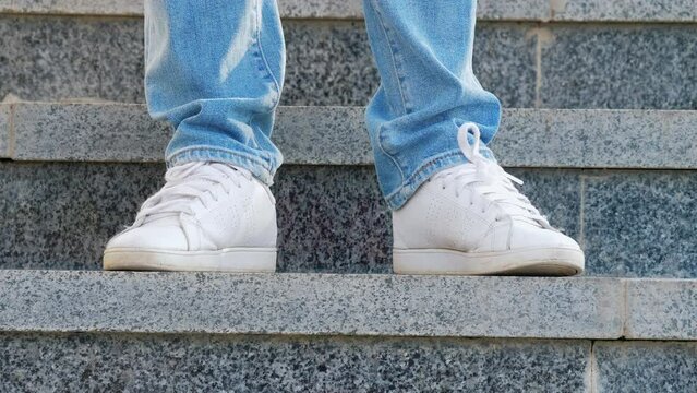 Legs Of Faceless Person In Jeans And White Sneakers Stand On Marble Steps. Girl In Walking Shoes Beats Rhythm Uniformly With Toe Of One Foot. Lonely Woman In Trainers Waiting For Someone At Street