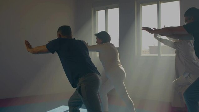Peaceful senior people practicing tai chi indoor. Medium shot of men and woman exercising with trainer in gym, bending toward legs, performing slow movements. Chinese martial arts, sport concept