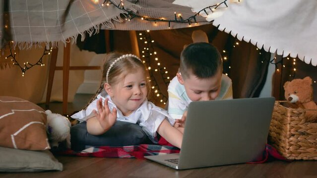 A little boy and girl communicate via video link via laptop while lying in a tent of blankets and pillows in the bedroom. Children