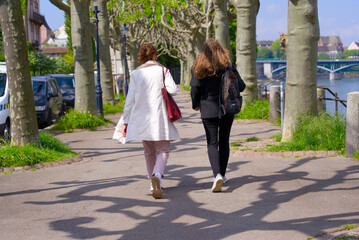 Two woman walking along a tree ally at border of Rhine River at City of Basel on a blue cloudy...