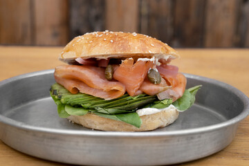 Gastronomy. Closeup view of a smoked salmon sandwich made with bagel bread, pink salmon filet, cream cheese, sliced avocado, capers, onion and arugula, in a metal dish on the wooden table.