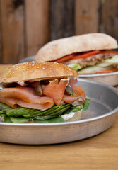 Sandwiches. Closeup view of smoked salmon bagel with cream cheese, sliced avocado, capers, onion, arugula and fried potatoes, and another sandwich in the background. 