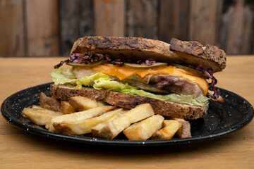 Sandwich. Closeup view of a bun with grilled chicken, lettuce, sliced cabbage, cucumber, cheddar cheese and fries, in a black dish on the wooden table.