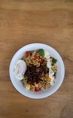 Top view of a Fusilli pasta salad with basil, crispy bacon, corn, cheese, tomato and a poached egg, in a white bowl on the wooden restaurant table.