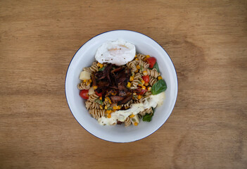 Top view of a Fusilli pasta salad with basil, crispy bacon, corn, cheese, tomato and a poached egg, in a white bowl on the wooden restaurant table. 