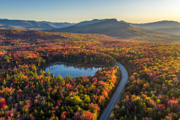 A road, highway through a forest with trees in fall colors of red, orange and yellow, lake,...