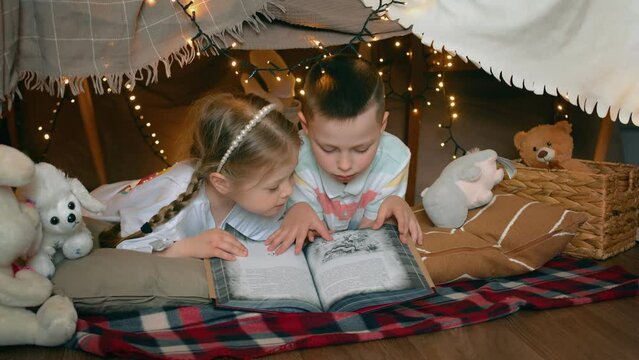 A little boy and girl are lying and reading a book in a tent of blankets and pillows in the bedroom. Children