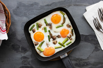 Fried eggs with vegetables (mushrooms and asparagus) in a small frying pan with cutlery, napkins and a wicker basket on dark grey stone background. Breakfast idea or concept. Top view, flat lay