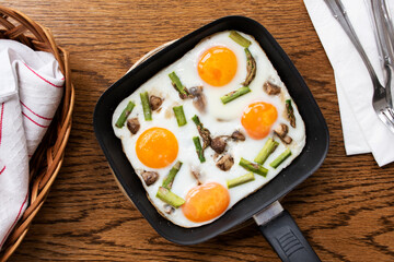 Fried eggs with vegetables (mushrooms and asparagus) in a small frying pan with cutlery, napkins and a wicker basket on wooden background. Breakfast idea or concept. Top view, flat lay