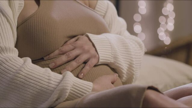 Pregnant woman touching big belly while sitting on bed. Close-up shot of womans hands. Unrecognizable expecting mother preparing for childbirth at home. Motherhood, pregnancy, childbirth concept