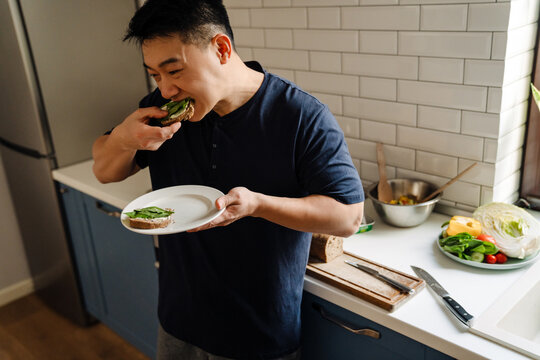 Adult asian man eating toats while standing on kozy kitchen