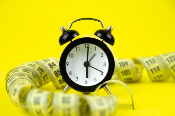 A black alarm clock with a white dial stands on a bright yellow background surrounded by a...