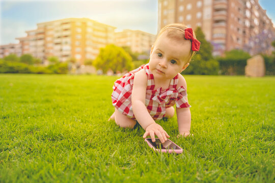 Curious little baby boy with blond curly hair dressed in light summer clothes sitting on lawn with fresh green grass sunny day playing on mobile phone, horizontal picture