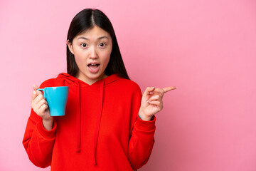 Young Chinese woman holding cup of coffee isolated on pink background surprised and pointing side