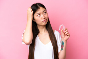 Young Chinese woman holding invisible braces isolated on pink background having doubts and with confuse face expression