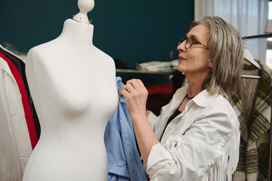 Elderly elegant Caucasian female fashion designer, trying on a stylish blue shirt on a mannequin in a fashion design workshop and tailoring atelier.