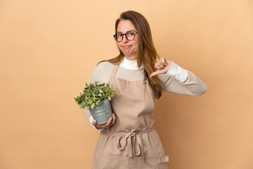 Middle age gardener woman holding a plant isolated on beige background showing thumb down with negative expression