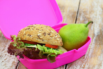Healthy school lunch box containing brown cheese roll with cheese, green lettuce, tomato and cucumber, and pear

