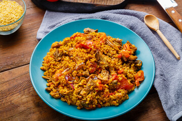 A large dish of bulgur with vegetables and mushrooms on a table on a napkin next to a wooden spoon.