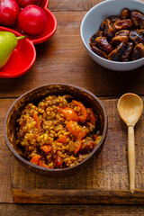 A plate of bulgur with vegetables and mushrooms on the table next to a wooden spoon and a cutting board.