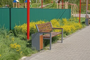 one brown wooden empty bench and a garbage urn stands on a gray sidewalk near green grass in a park outdoors