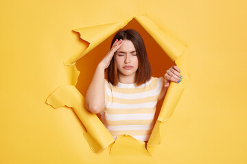 Indoor shot of tired exhausted young adult brunette woman stands in torn paper hole, suffering headache, touching head, looking through breakthrough of yellow background.