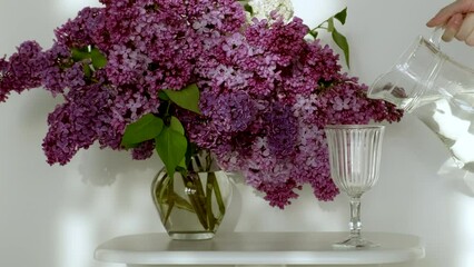 Mineral water in wineglass. Unrecognizable person pouring clean water in glass. Pour pure water into glass from jug at white background, vase with a lilac flowers near on table. Close up, slow motion