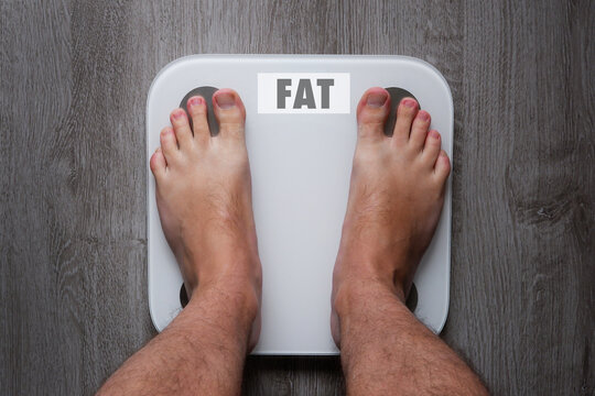 Top View Of The Feet Of A Barefoot Man Standing On Smart Scales, On Whose Balance It Is Written Fat