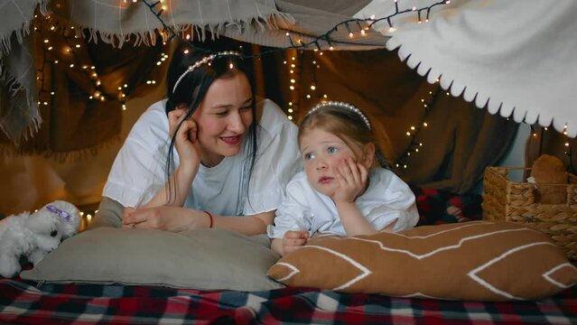 Mom and little daughter communicate while lying in a tent made of blankets and pillows in the bedroom. A family