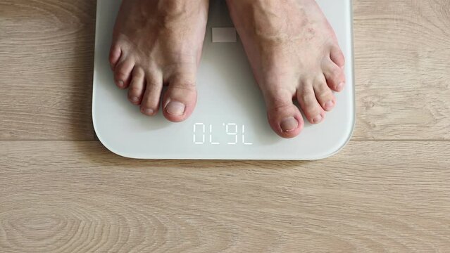 close up of Man walking on scales measure weight. male wal checking BMI weight loss. human barefoot measuring body fat overweight. Guy legs step on bathroom scale. top view.