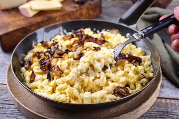 Traditionelle Schweizer Käsespätzle mit gerösteten Zwiebeln serviert als close-up in einer Eisenpfanne auf einem rustikalen Holzbrett in einer Berghütte