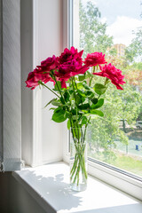 A bouquet of red roses in a transparent glass vase, which stands on the windowsill.