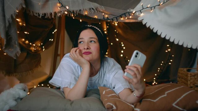 A young woman takes a selfie on her smartphone while lying in a tent of blankets and pillows in the bedroom. Leisure