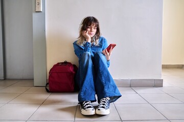 Sad young female student with backpack smartphone sitting on the floor