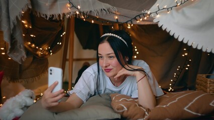 A young woman takes a selfie on her smartphone while lying in a tent of blankets and pillows in the bedroom. Leisure