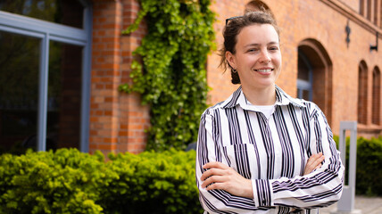 Young calm smiling freelance woman in white striped shirt looking at camera, standing outdoors on red brick building office, big window, green bush background. Portrait of happy female IT specialist