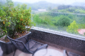 View from the window covered with raindrops on a summer landscape and a balcony with a flower bed a...