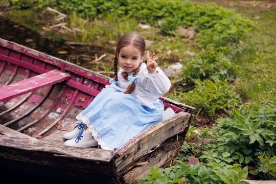 Little 5-eyar-old girl sitting in old wooden pink boat in village. Little baby girl of 5-years-old in retro vintage dress serious and thoughtful