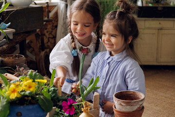 Two litlle sisters planting plant sets on wooden terrace. Two little baby girls of 3-years-old and 5-years-old in retro vintage dresses digging with a shovel soil and smiling. Child  fun and rest