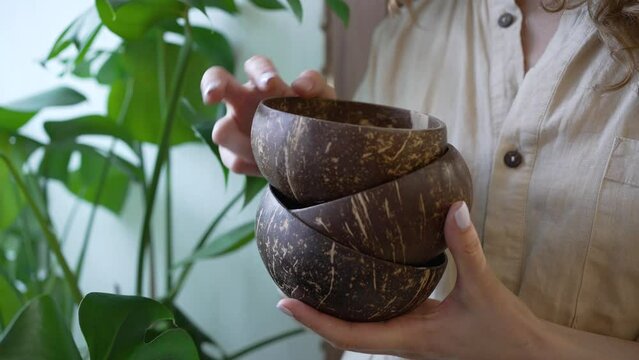 Young woman holds small brown empty bowls in hands with white manicure against green plant. Woman overlooks new dishware for cafe closeup