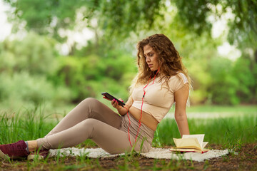 Naklejka na ściany i meble Image of positive caucasian woman lying outdoors in park on a grass and using mobile phone while listening music.
