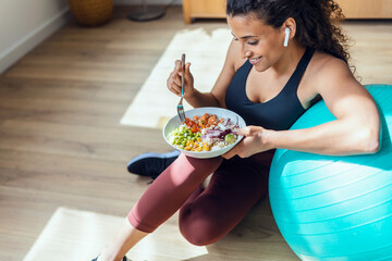 Sporty young woman eating healthy while listening to music sitting on the floor at home. - obrazy, fototapety, plakaty