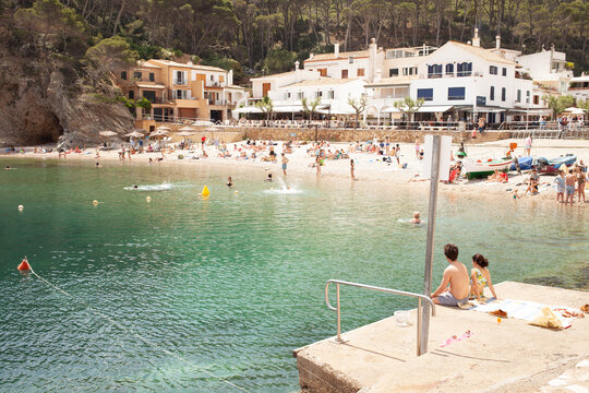 Fisherman's town on Mediterranean sea cost. People enjoying sun, sunbathing, swimming in transparent blue sea. Sandy beach with clear, shallow water, with kayak & SUP rentals & seafront eateries.