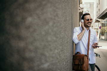 Casual dressed young business man standing by the wall listening to music, holding hands on wireless headphones