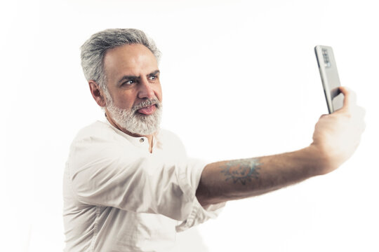 Medium studio shot on white background of an old good-looking funny man holding a mobilephone, making a selfie, sticking his tongue out. High quality photo